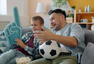 Football fans father and child are watching match on TV at home holding foam hand and ball. Spending time with family, cheering and sports concept.