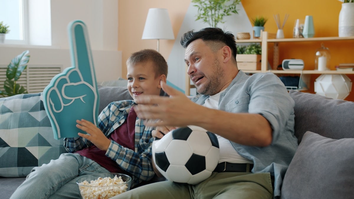 Football fans father and child are watching match on TV at home holding foam hand and ball. Spending time with family, cheering and sports concept.